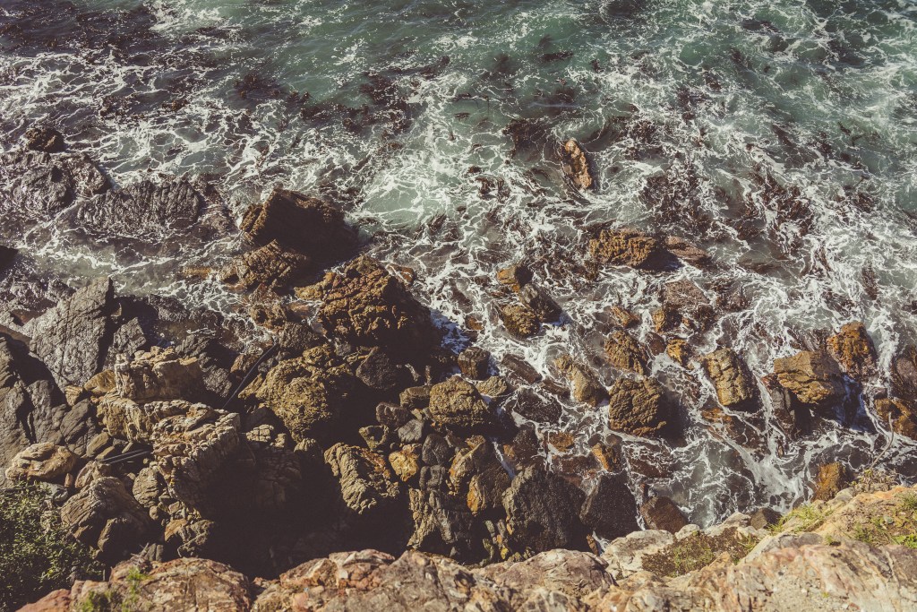 Aerial view of rocky shoreline with waves crashing against the rocks and sea foam visible.